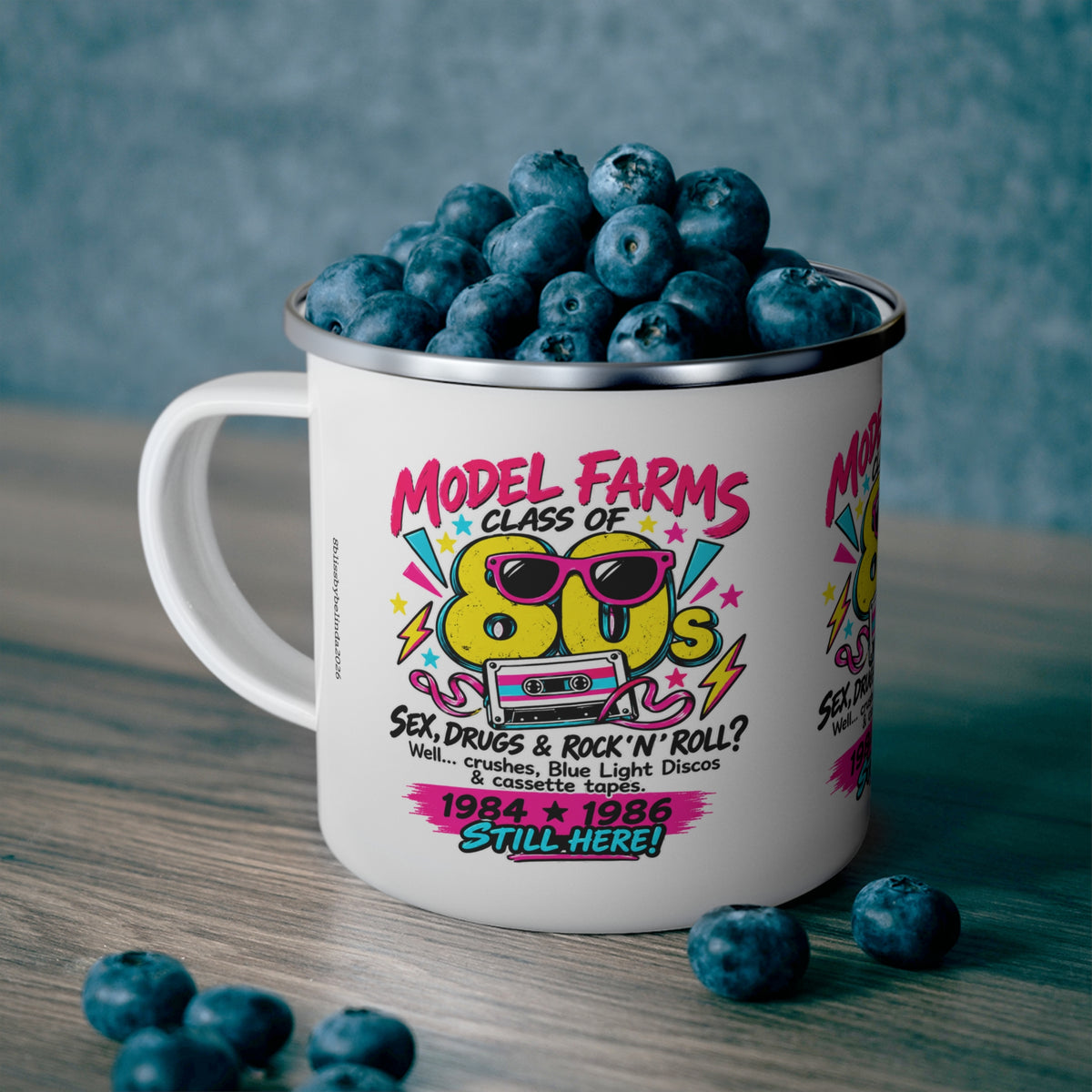 White enamel camping mug with colorful retro cassette graphic and wide handle, filled with blueberries